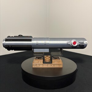 May include: A silver lightsaber prop with black and red accents, resting on a black base with a decorative brown stand. The lightsaber features a detailed design with a red button and a black handle.