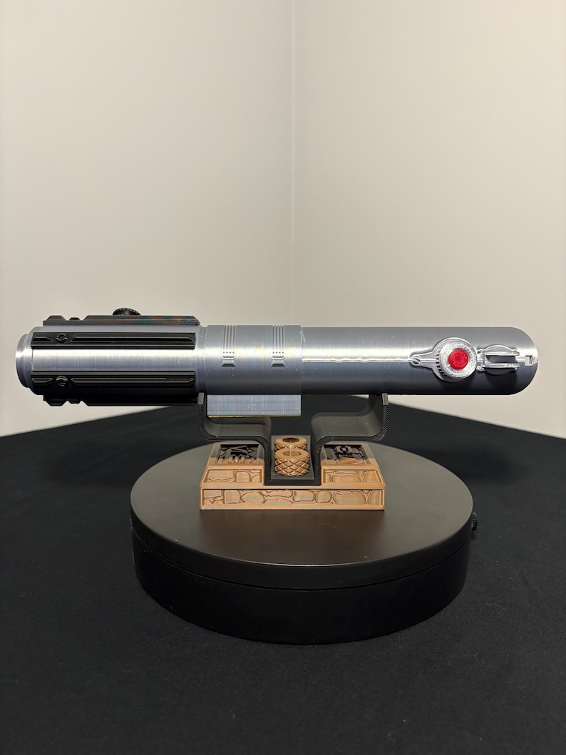 May include: A silver lightsaber prop with black and red accents, resting on a black base with a decorative brown stand. The lightsaber features a detailed design with a red button and a black handle.