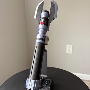 May include: A futuristic-looking prop lightsaber with a black and blue handle, a red button, and silver blades. The lightsaber is mounted on a black stand, showcasing its unique design and details. The overall aesthetic suggests a science fiction theme.