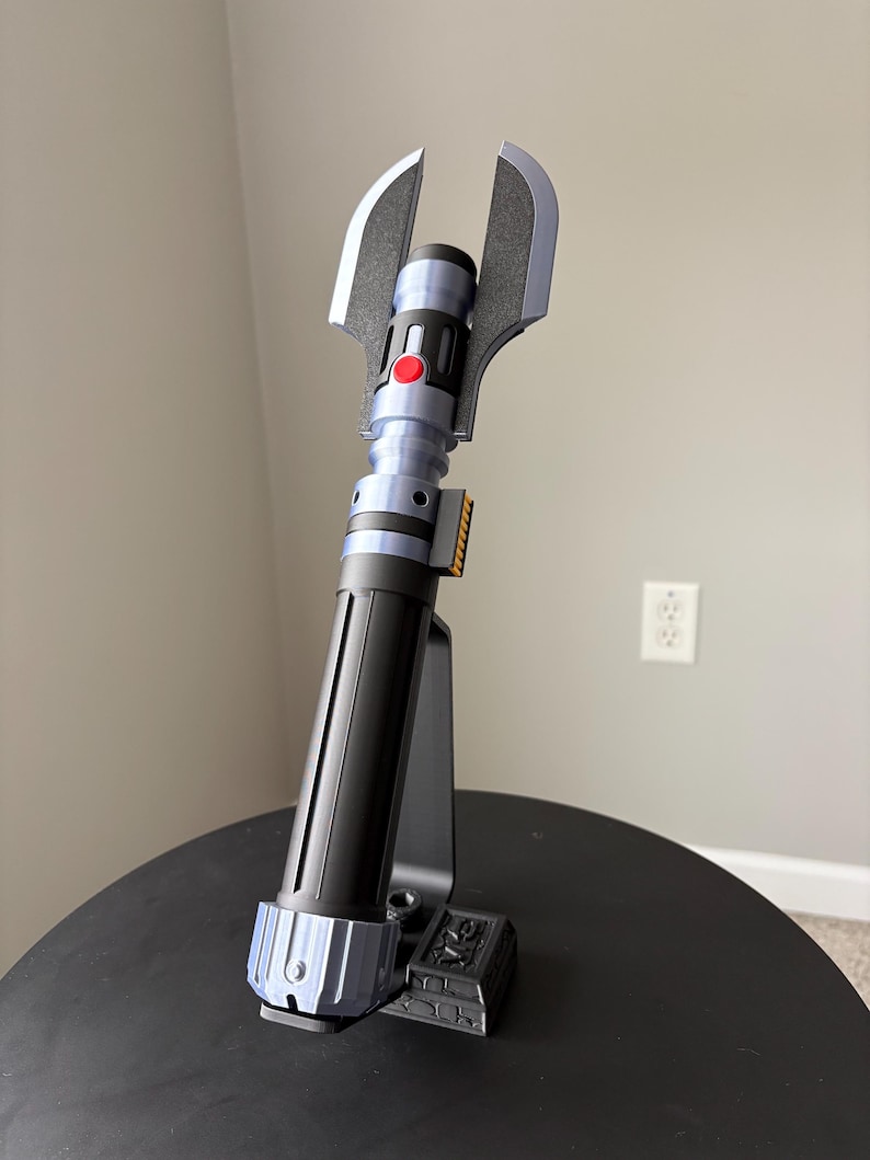 May include: A futuristic-looking prop lightsaber with a black and blue handle, a red button, and silver blades. The lightsaber is mounted on a black stand, showcasing its unique design and details. The overall aesthetic suggests a science fiction theme.