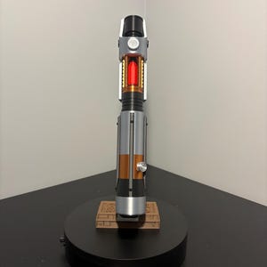 May include: A detailed lightsaber prop with a black, silver, and copper handle. The center features a red, glowing crystal. The lightsaber rests on a black base with a brown, rectangular platform.