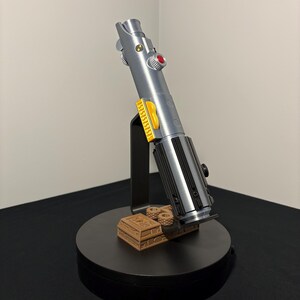 May include: A silver and black lightsaber prop with yellow and red accents, displayed on a black stand with a brown base. The prop is a replica of a lightsaber from a science fiction franchise.