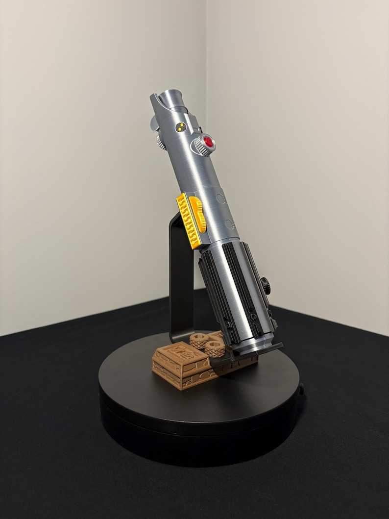 May include: A silver and black lightsaber prop with yellow and red accents, displayed on a black stand with a brown base. The prop is a replica of a lightsaber from a science fiction franchise.