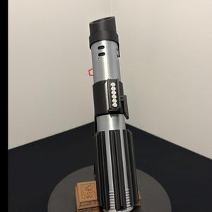 May include: A replica lightsaber, featuring a black and silver design with a ribbed handle and a cylindrical emitter. The prop is displayed on a black stand with a brown base, showcasing a detailed replica of a fictional weapon.
