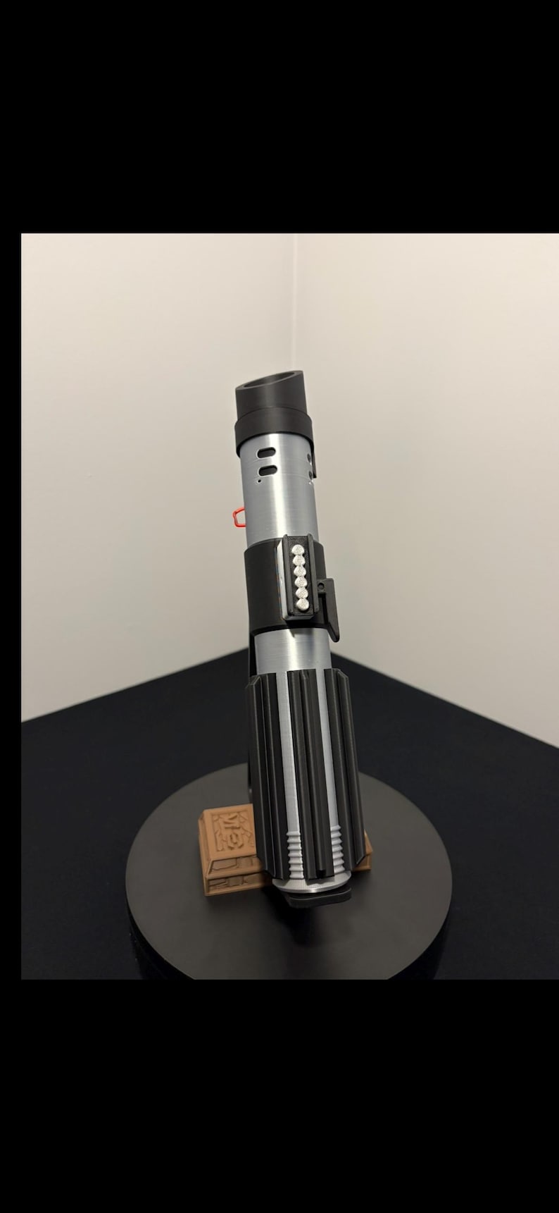 May include: A replica lightsaber, featuring a black and silver design with a ribbed handle and a cylindrical emitter. The prop is displayed on a black stand with a brown base, showcasing a detailed replica of a fictional weapon.