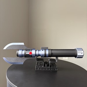 May include: A black and blue lightsaber with a unique blade design, featuring a red button and silver accents. The lightsaber is displayed on a black stand, showcasing its intricate details and futuristic aesthetic.