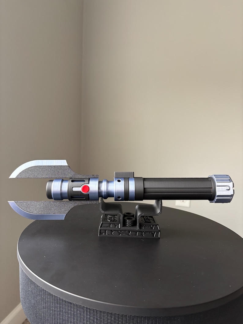 May include: A black and blue lightsaber with a unique blade design, featuring a red button and silver accents. The lightsaber is displayed on a black stand, showcasing its intricate details and futuristic aesthetic.