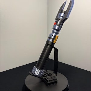 May include: A black and silver lightsaber prop with a red button and yellow accents, displayed on a black stand. The lightsaber has a detailed design and is angled upwards, showcasing its blade-like features.
