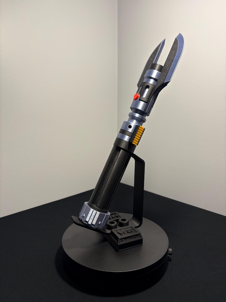 May include: A black and silver lightsaber prop with a red button and yellow accents, displayed on a black stand. The lightsaber has a detailed design and is angled upwards, showcasing its blade-like features.