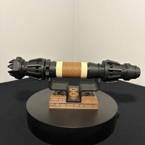 May include: A black and brown lightsaber prop with a brown leather-wrapped grip and tan accents. The prop is displayed on a black stand with a detailed, brown base. The lightsaber is a detailed replica.