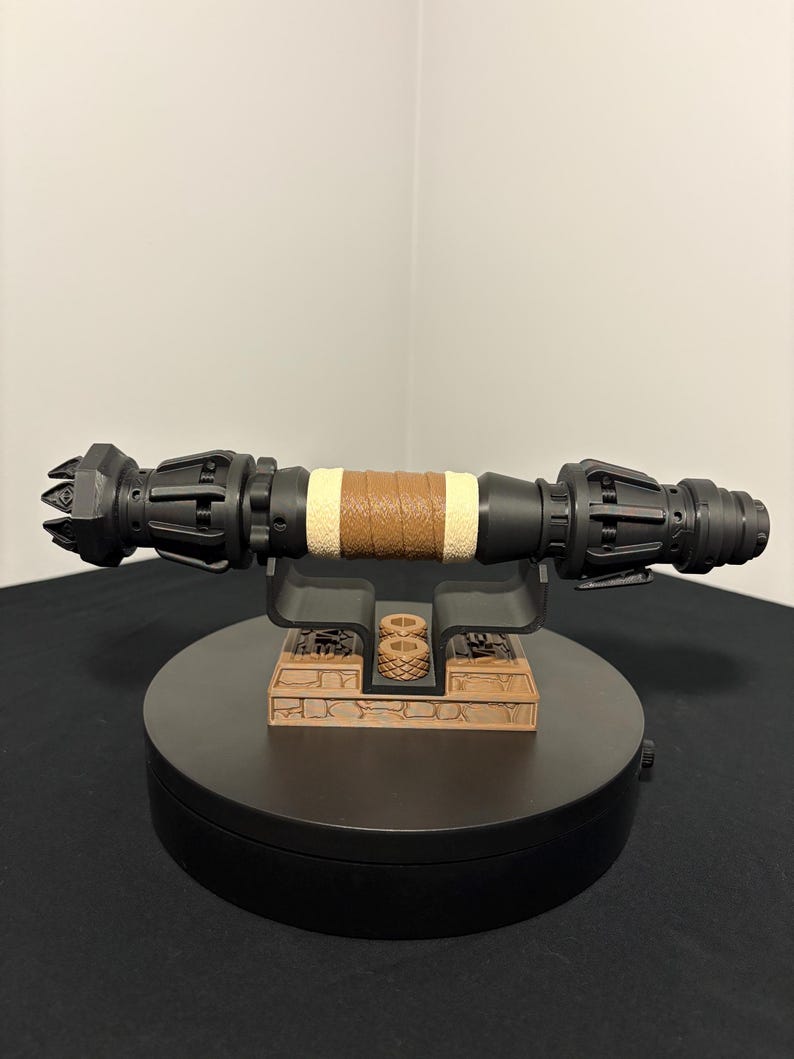 May include: A black and brown lightsaber prop with a brown leather-wrapped grip and tan accents. The prop is displayed on a black stand with a detailed, brown base. The lightsaber is a detailed replica.
