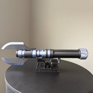 May include: A black and blue lightsaber prop with a unique blade design. The prop is mounted on a black stand, showcasing its intricate details. The lightsaber features a cylindrical handle and a distinctive blade shape.