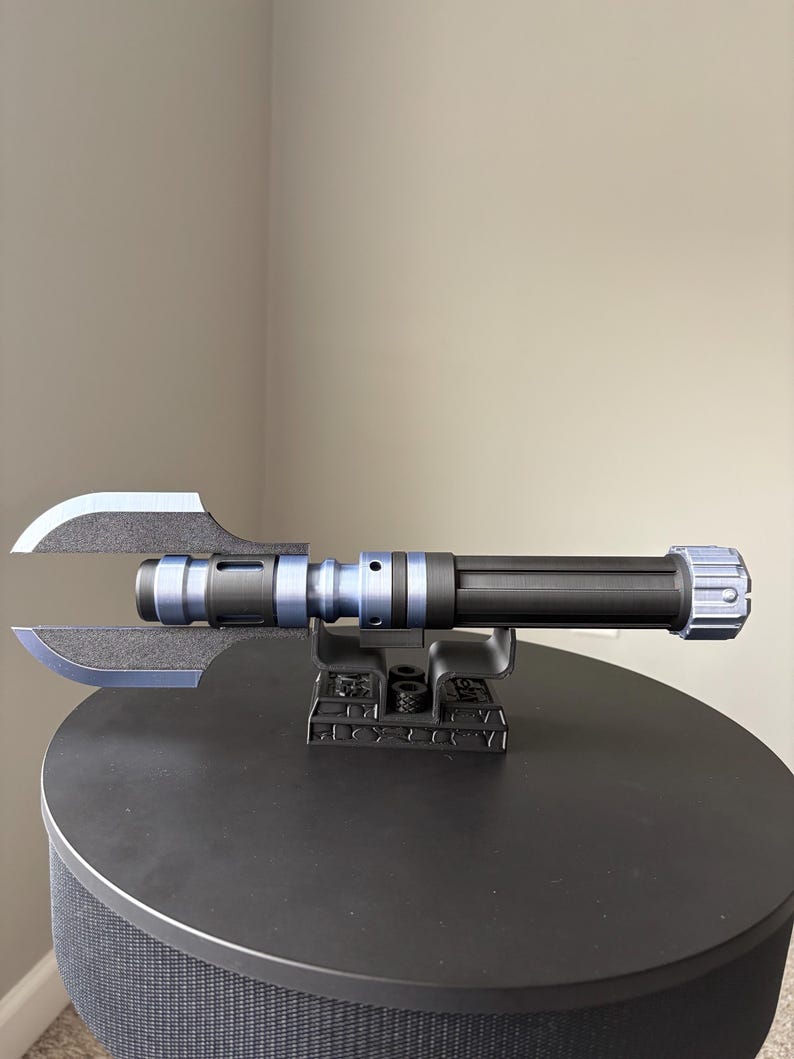 May include: A black and blue lightsaber prop with a unique blade design. The prop is mounted on a black stand, showcasing its intricate details. The lightsaber features a cylindrical handle and a distinctive blade shape.