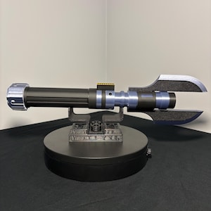 May include: A black and blue lightsaber prop with a unique blade design, resting on a black display stand. The lightsaber features silver and blue accents, and is set against a neutral background.