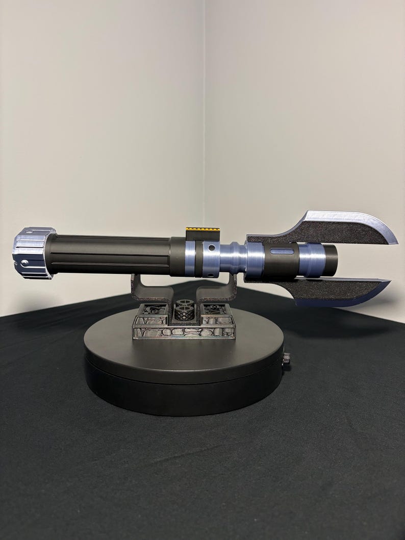 May include: A black and blue lightsaber prop with a unique blade design, resting on a black display stand. The lightsaber features silver and blue accents, and is set against a neutral background.