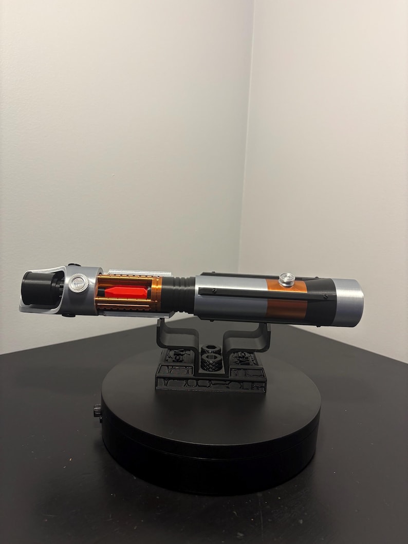 May include: A detailed replica lightsaber with a black, silver, and orange color scheme. The hilt features intricate detailing and a red internal component. It is displayed on a black stand, showcasing its design.