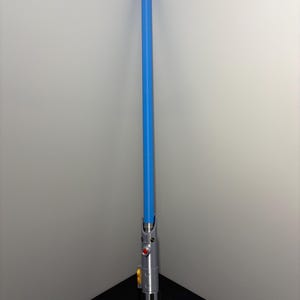 May include: A blue lightsaber prop with a silver handle. The handle features red and yellow buttons, and a black grip. The blue blade extends upwards, creating a striking contrast against the silver and black.