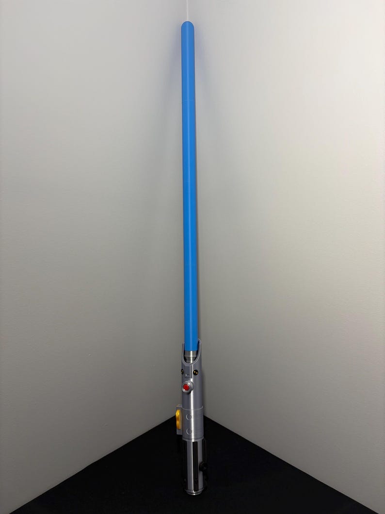 May include: A blue lightsaber prop with a silver handle. The handle features red and yellow buttons, and a black grip. The blue blade extends upwards, creating a striking contrast against the silver and black.