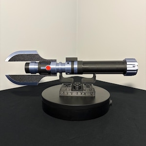 May include: A black and silver lightsaber prop with a unique blade design. The hilt features blue and black accents, a red button, and a silver end cap. The prop is displayed on a black rotating stand.