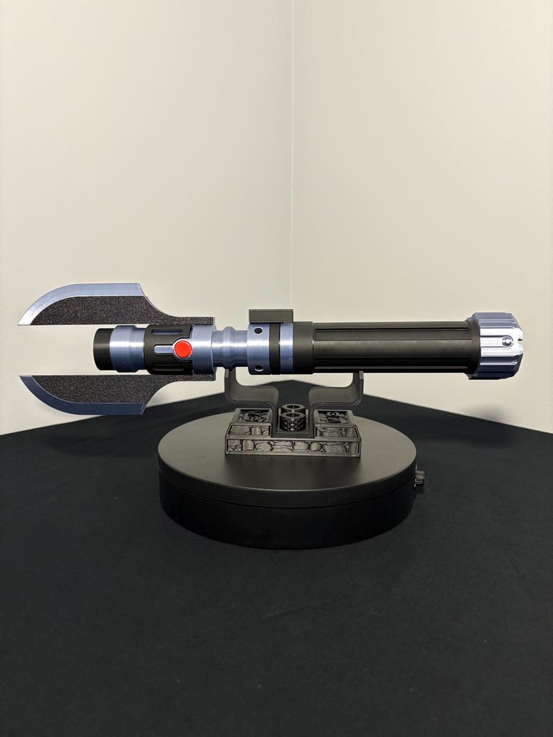 May include: A black and silver lightsaber prop with a unique blade design. The hilt features blue and black accents, a red button, and a silver end cap. The prop is displayed on a black rotating stand.