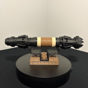May include: A black and brown lightsaber prop with a tan leather-wrapped grip. The prop is displayed on a black rotating stand with a brown base. The lightsaber has intricate details and a futuristic design.