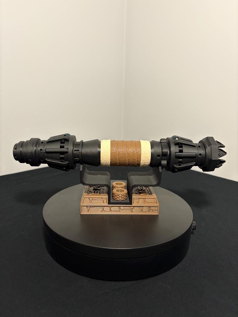 May include: A black and brown lightsaber prop with a tan leather-wrapped grip. The prop is displayed on a black rotating stand with a brown base. The lightsaber has intricate details and a futuristic design.