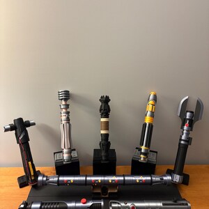 May include: A collection of replica lightsabers in various designs and colors, displayed on wooden and black stands. The lightsabers feature metallic silver, black, yellow, and red accents. Some have unique handle designs, and all are presented against a neutral gray background.