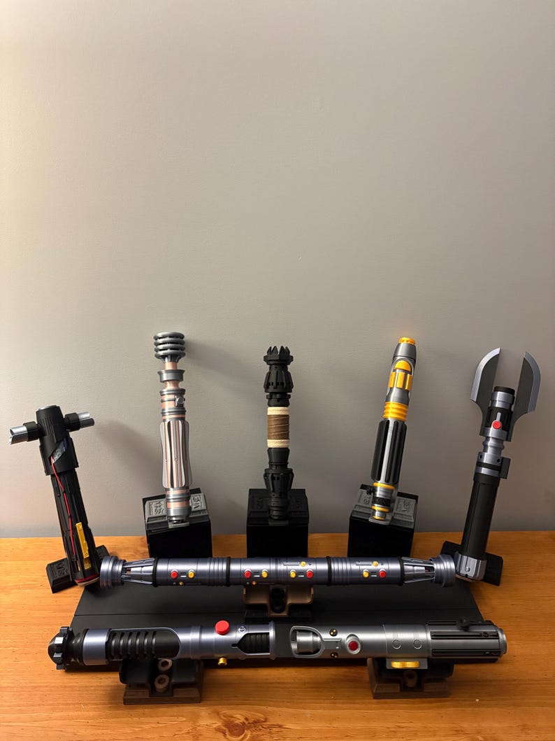 May include: A collection of replica lightsabers in various designs and colors, displayed on wooden and black stands. The lightsabers feature metallic silver, black, yellow, and red accents. Some have unique handle designs, and all are presented against a neutral gray background.