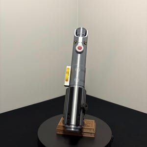 May include: A silver and black lightsaber replica with red and yellow accents, displayed on a black circular stand. The lightsaber has a detailed design and is mounted on a brown rectangular base.