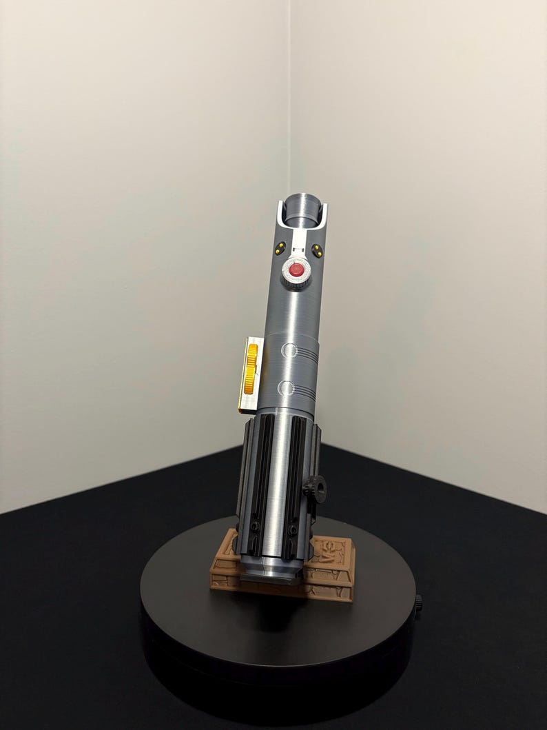 May include: A silver and black lightsaber replica with red and yellow accents, displayed on a black circular stand. The lightsaber has a detailed design and is mounted on a brown rectangular base.