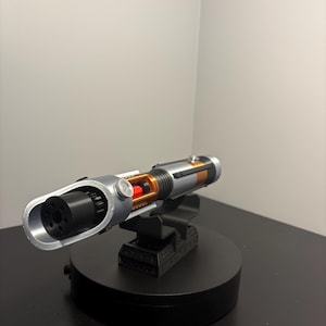 May include: A silver and black lightsaber prop with orange and red accents, mounted on a black display stand. The prop features a detailed design with visible internal components and a cylindrical handle. The stand is on a black surface.