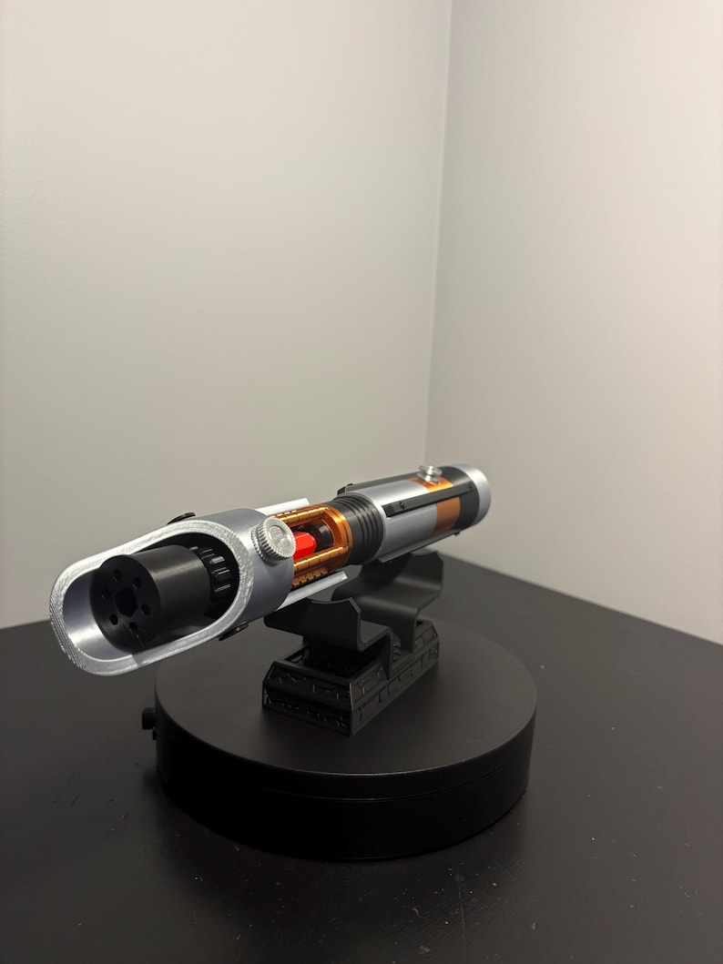 May include: A silver and black lightsaber prop with orange and red accents, mounted on a black display stand. The prop features a detailed design with visible internal components and a cylindrical handle. The stand is on a black surface.