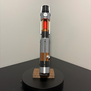 May include: A detailed lightsaber prop with a black, silver, and copper handle. The center features a red, glowing crystal. The lightsaber rests on a black base with a brown, rectangular platform.