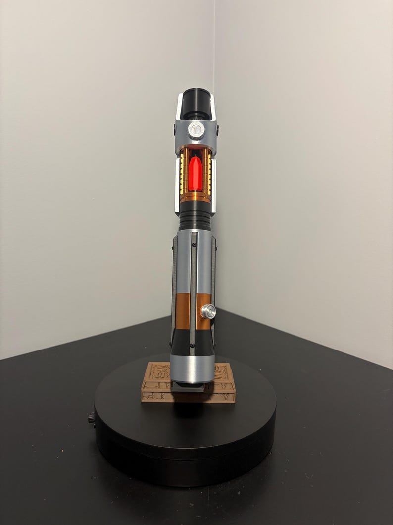 May include: A detailed lightsaber prop with a black, silver, and copper handle. The center features a red, glowing crystal. The lightsaber rests on a black base with a brown, rectangular platform.