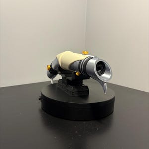 May include: A futuristic-looking, silver and black device with a cream-colored grip and gold accents. The device is mounted on a black base, suggesting a display or collectible item. The design is sleek and detailed.