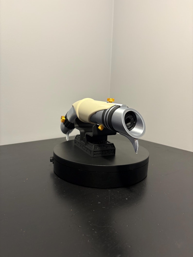 May include: A futuristic-looking, silver and black device with a cream-colored grip and gold accents. The device is mounted on a black base, suggesting a display or collectible item. The design is sleek and detailed.