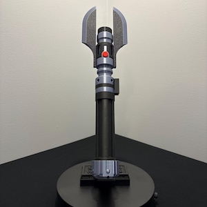 May include: A black and gray lightsaber prop with a silver blade. The hilt features a red button and a unique blade guard design. The lightsaber is displayed on a black circular stand.