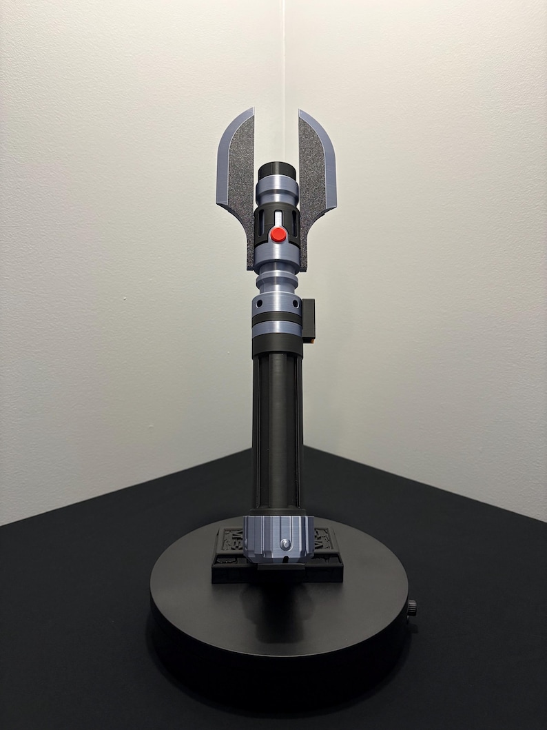 May include: A black and gray lightsaber prop with a silver blade. The hilt features a red button and a unique blade guard design. The lightsaber is displayed on a black circular stand.