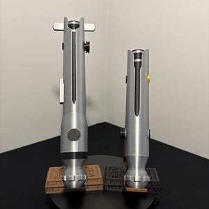 May include: Two silver-colored lightsabers, each with a black central blade, are displayed on a black stand. The lightsabers have detailed handles and are mounted on brown and black rectangular bases. The background is a plain white wall.