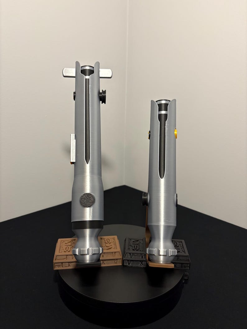 May include: Two silver-colored lightsabers, each with a black central blade, are displayed on a black stand. The lightsabers have detailed handles and are mounted on brown and black rectangular bases. The background is a plain white wall.