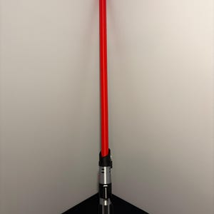 May include: A red lightsaber with a black and silver hilt. The lightsaber is standing upright in the corner of a room. The hilt has a detailed design with various buttons and a ribbed grip.