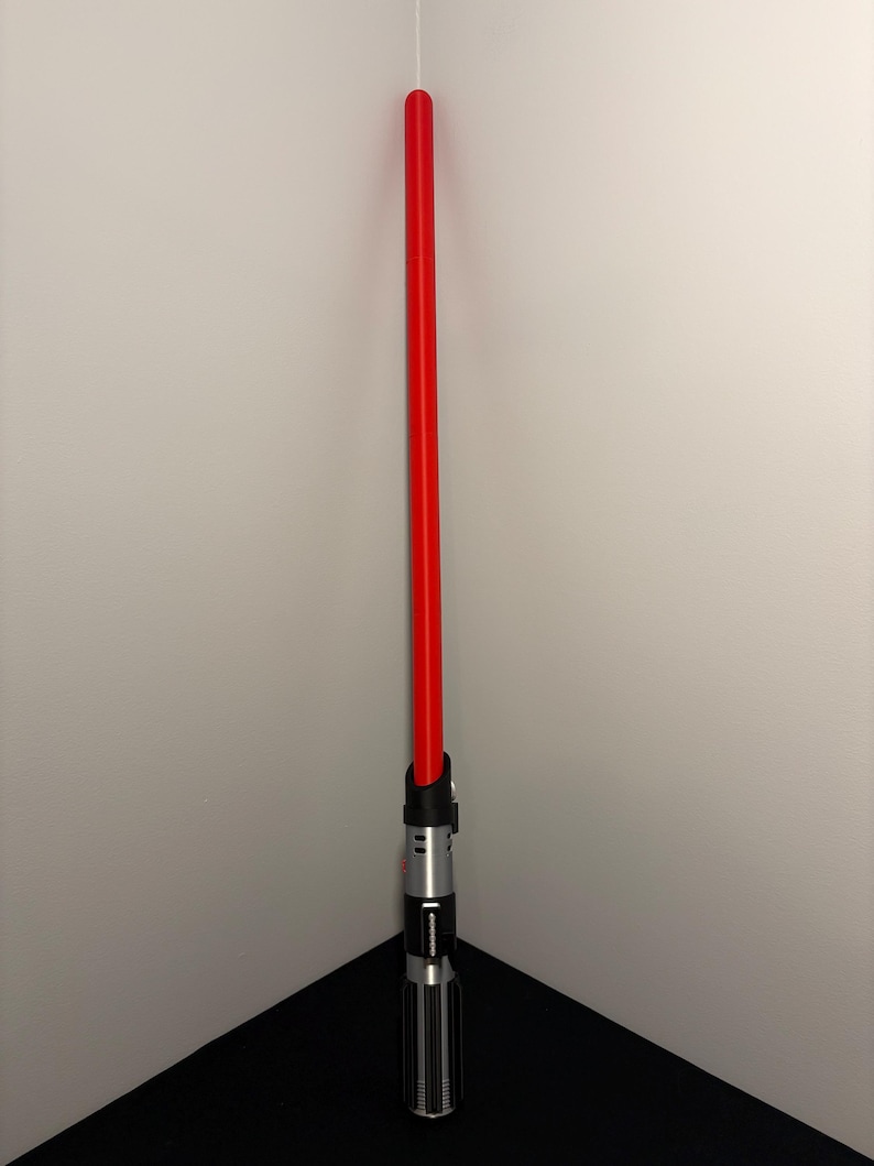 May include: A red lightsaber with a black and silver hilt. The lightsaber is standing upright in the corner of a room. The hilt has a detailed design with various buttons and a ribbed grip.