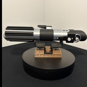 May include: A detailed replica lightsaber with a black and silver handle, resting on a black display stand. The lightsaber features intricate detailing and a cylindrical design, showcasing a classic sci-fi aesthetic.