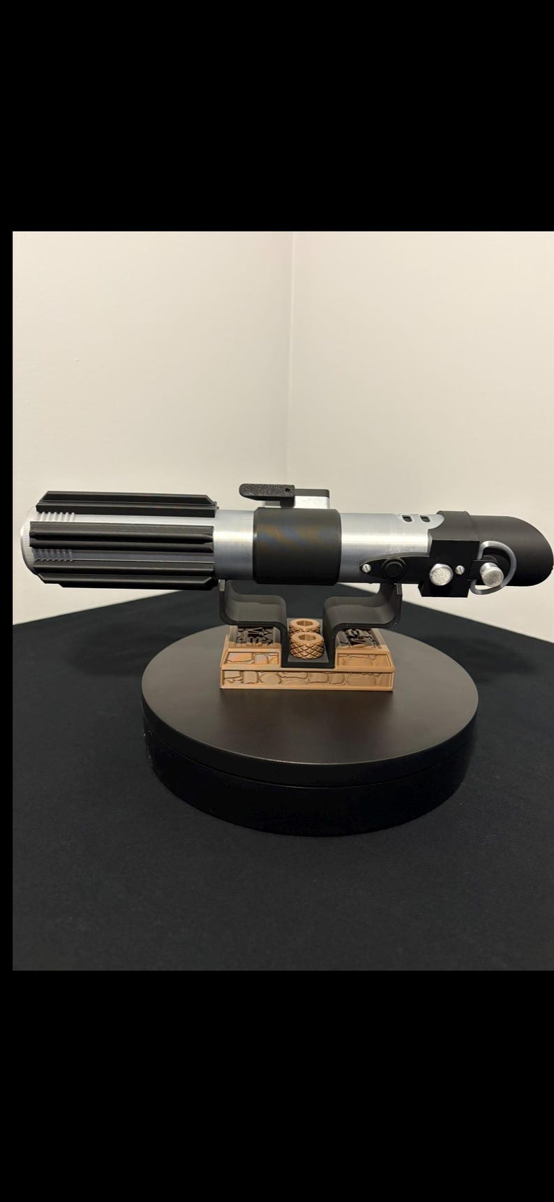 May include: A detailed replica lightsaber with a black and silver handle, resting on a black display stand. The lightsaber features intricate detailing and a cylindrical design, showcasing a classic sci-fi aesthetic.