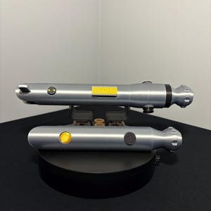 May include: Two silver-colored toy lightsabers, one above the other, are displayed on a black surface. Each has a cylindrical body with a button and a small yellow rectangular detail. The lightsabers are designed with a futuristic aesthetic.