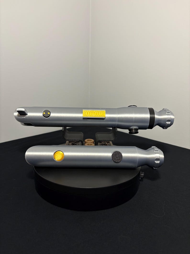 May include: Two silver-colored toy lightsabers, one above the other, are displayed on a black surface. Each has a cylindrical body with a button and a small yellow rectangular detail. The lightsabers are designed with a futuristic aesthetic.