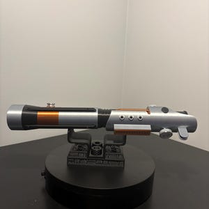 May include: A silver and black lightsaber prop with copper-colored accents, resting on a black display stand. The prop features a cylindrical design with detailed mechanical elements, including circular buttons and a control knob.
