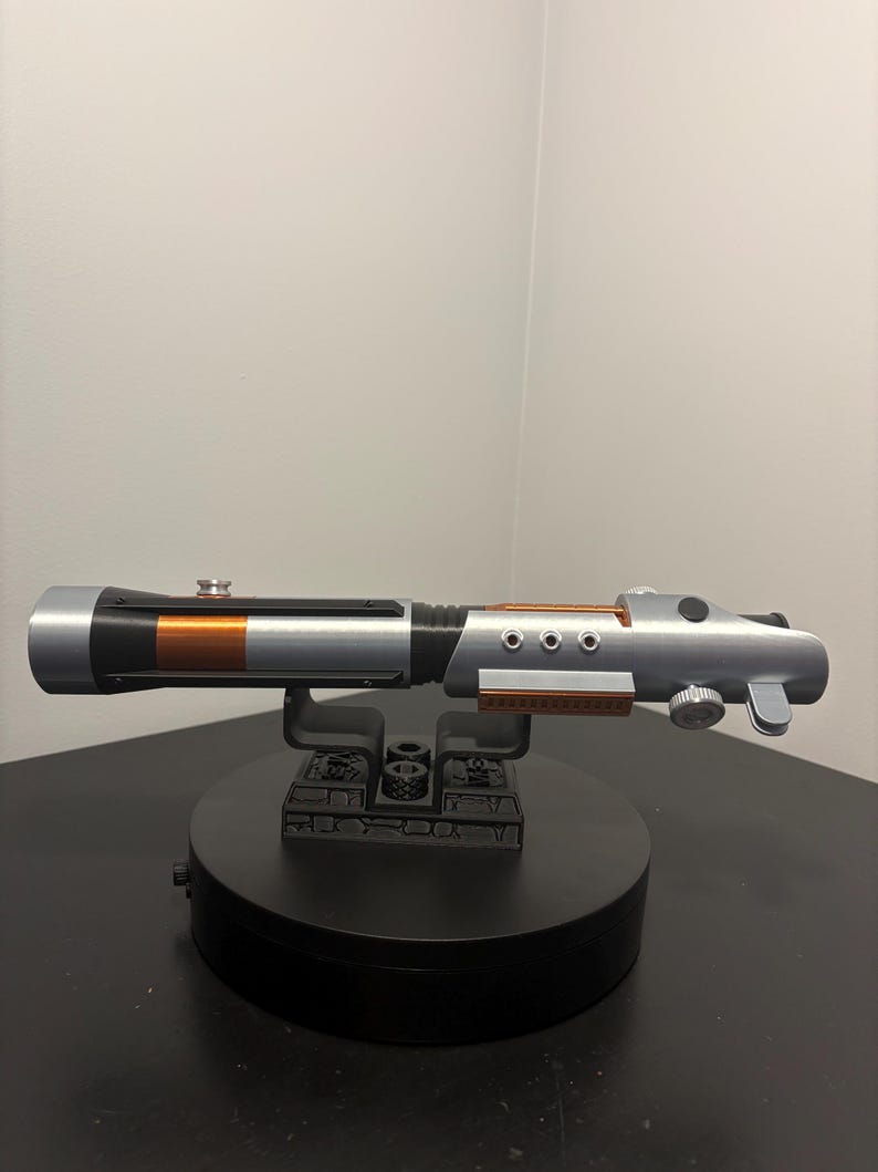 May include: A silver and black lightsaber prop with copper-colored accents, resting on a black display stand. The prop features a cylindrical design with detailed mechanical elements, including circular buttons and a control knob.