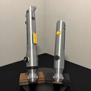 May include: Two silver lightsabers with yellow and black accents, standing on brown and black bases. The lightsabers have detailed handles and are displayed on a black circular stand. The background is a plain white wall.
