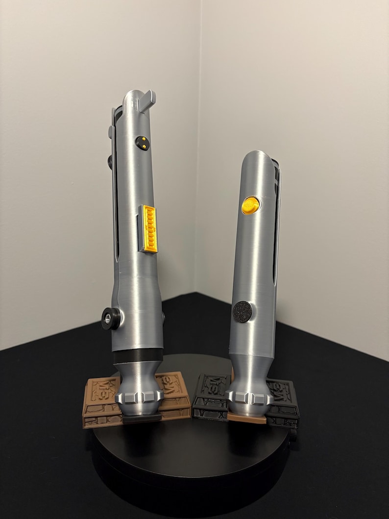 May include: Two silver lightsabers with yellow and black accents, standing on brown and black bases. The lightsabers have detailed handles and are displayed on a black circular stand. The background is a plain white wall.